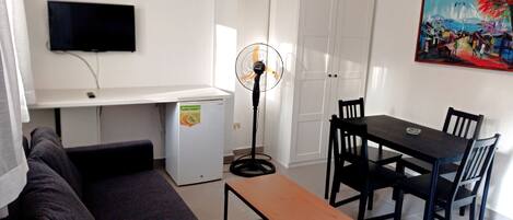 Een woonkamer met een bank, een kleine tafel, een televisie, een ventilator en een eethoek met stoelen en een tafel.