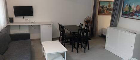 A compact hotel room with a bed, a sofa, a small table, a desk with a TV, a fan, and a painting on the wall.