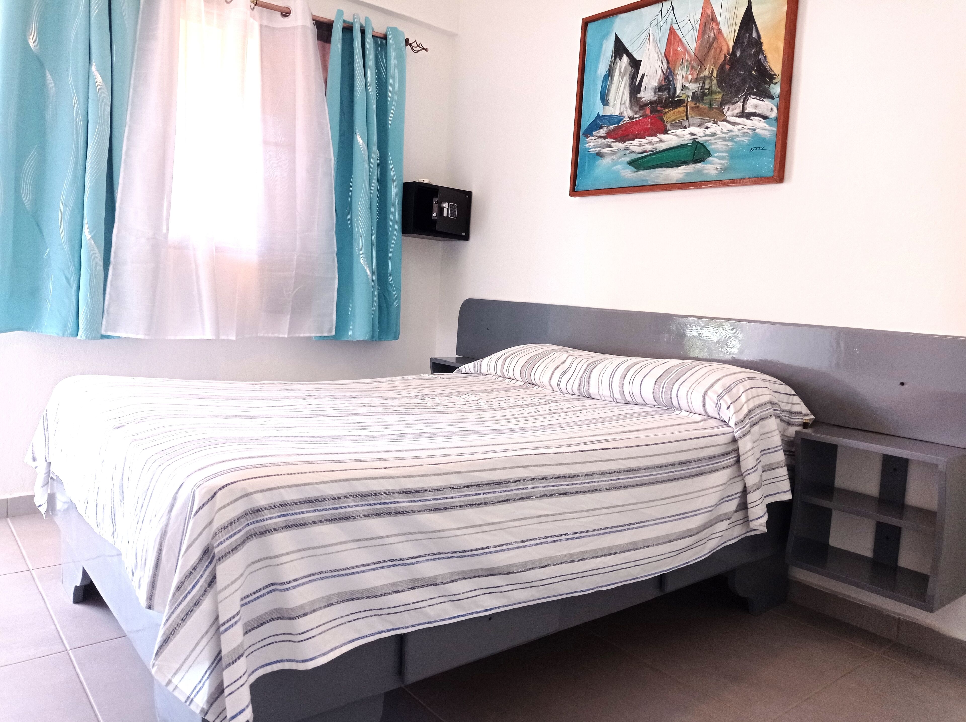 A bed with a striped bedspread, a bedside table, a nightstand, and a painting of sailboats on the wall.