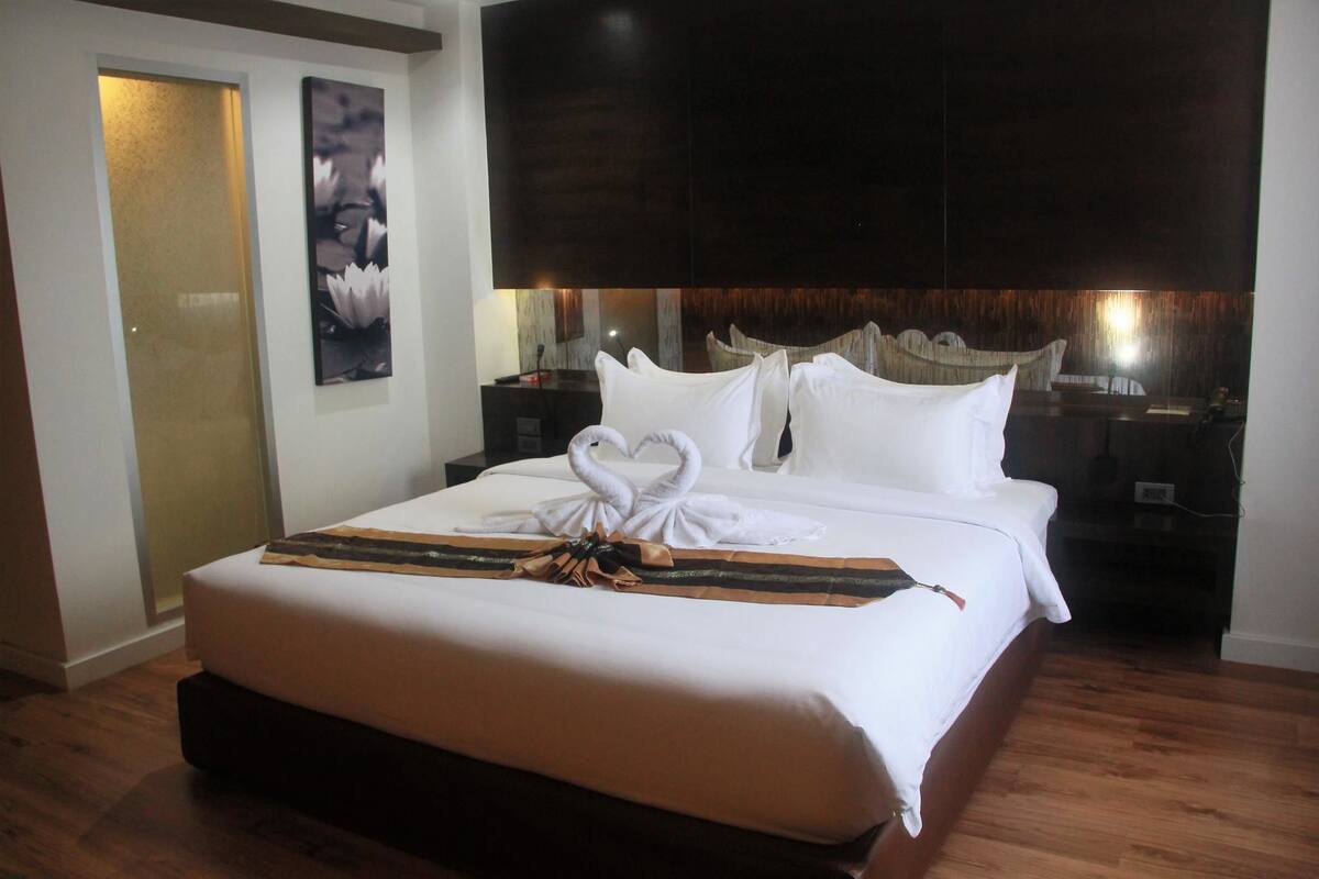 A hotel room with a large bed, a flat-screen TV, a wooden headboard, and a nightstand with a lamp.