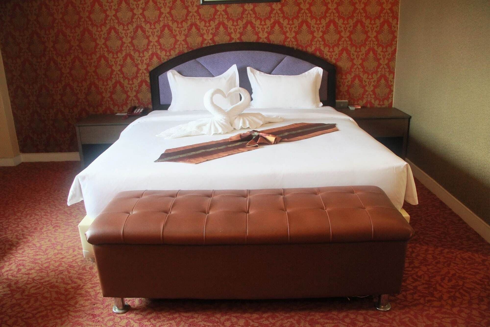 A neatly made bed with a brown upholstered bench in front, a decorative swan pillow, and a folded tie on the bed.