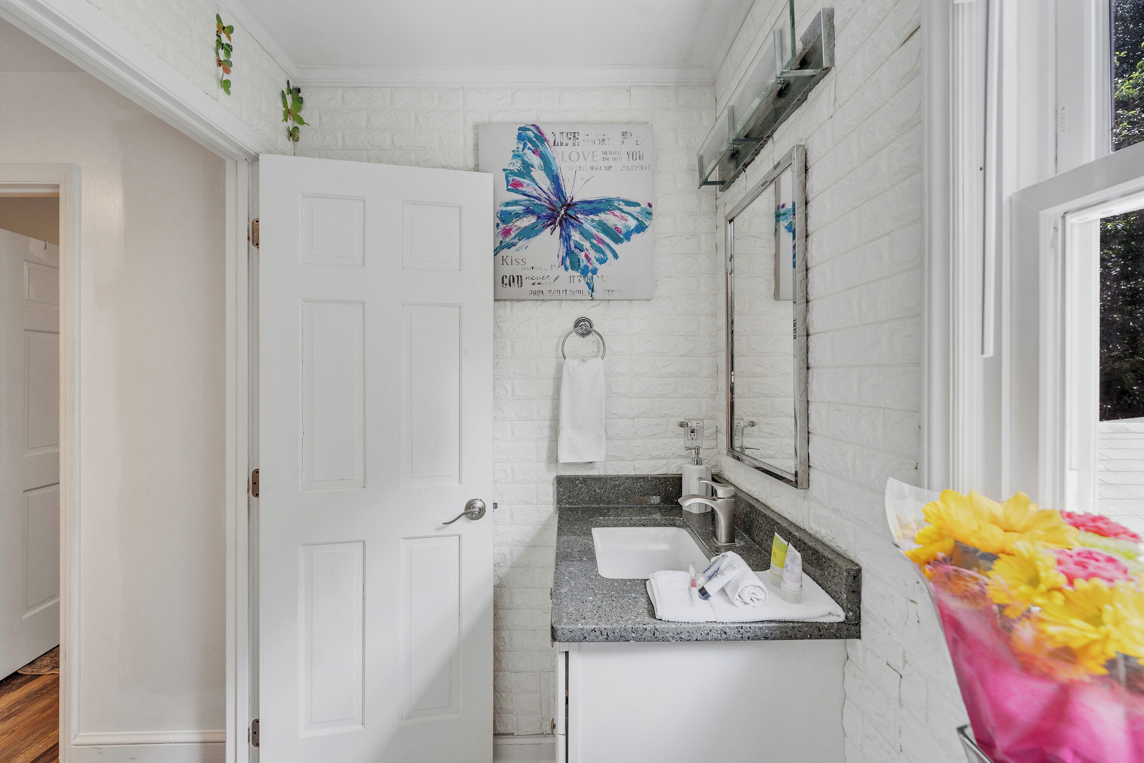 Une salle de bain avec une porte blanche, un lavabo avec un miroir et une œuvre d’art représentant un papillon accrochée au mur.