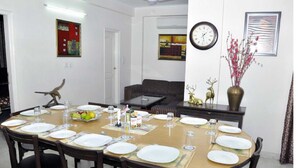 A dining area with a round table set for six, surrounded by chairs. There is a sofa in the background, a clock on the wall, and a vase with flowers on a side table. - Mehra Residency at the Airport (New Delhi)