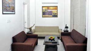 A living room with a beige sofa, two brown armchairs, a coffee table with fruit, and artwork on the walls. - Mehra Residency at the Airport (New Delhi)