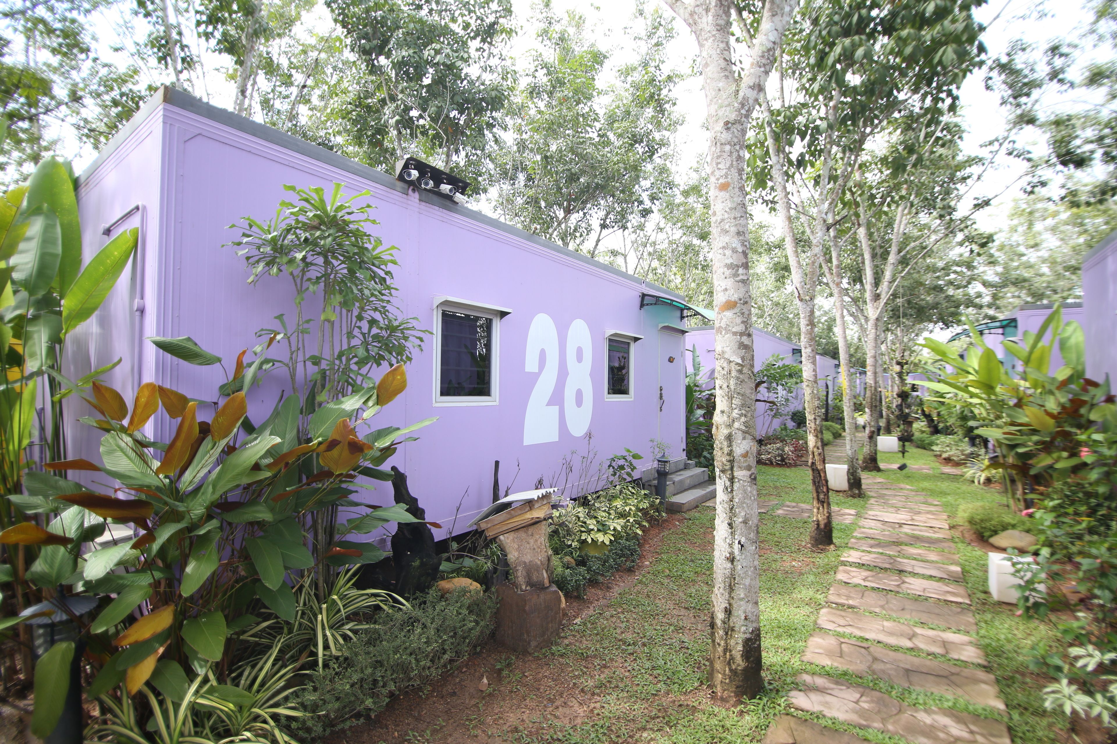 A row of purple mobile homes numbered 28, surrounded by lush greenery and a stone pathway.