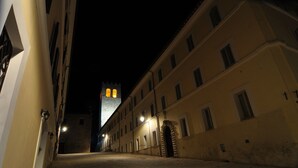 Una calle estrecha flanqueada por edificios, una torre con ventanas iluminadas a lo lejos, y un camino empedrado.