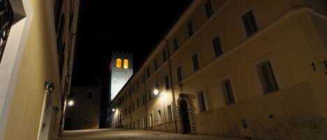 A narrow street with buildings on either side, a tower with illuminated windows in the distance, and a cobblestone path.