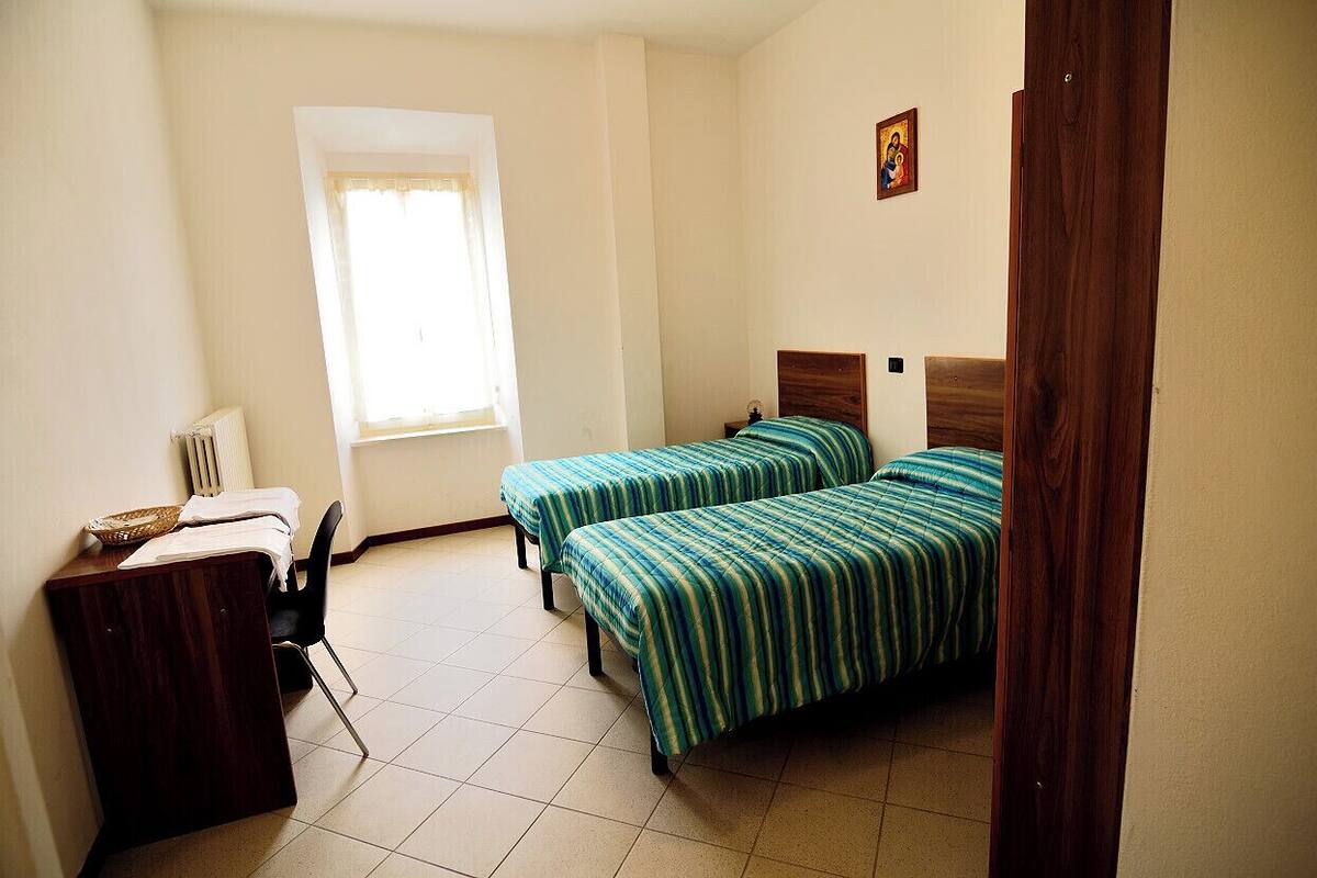 A room with two single beds, a desk with a chair, a radiator, and a window with curtains.