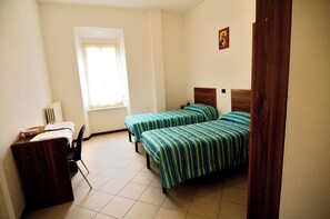 A room with two single beds, a desk with a chair, a radiator, and a window with curtains.