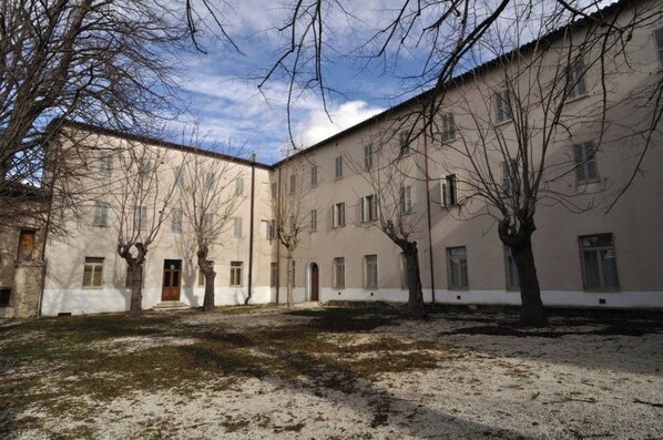 A courtyard with a building featuring multiple windows and a central door, surrounded by trees and a grassy area.