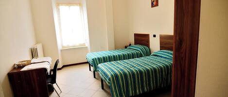 A room with two single beds, a desk with a chair, a radiator, and a window with curtains.