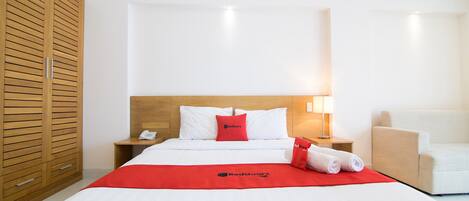 A hotel room with a bed, a wooden wardrobe, a bedside table with a lamp, and a white armchair.