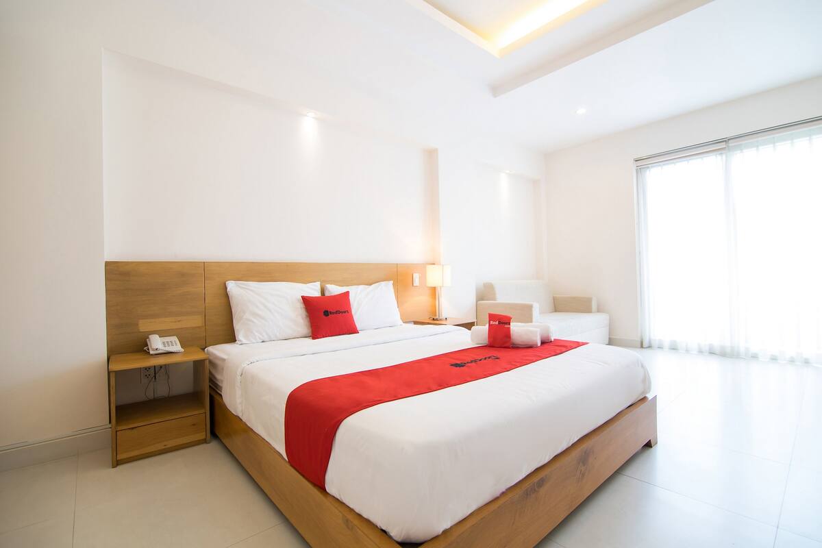 Ein modernes Hotelzimmer mit einem großen Bett, einem Nachttisch mit Telefon, einer Lampe und einem Fenster mit durchsichtigen Vorhängen.