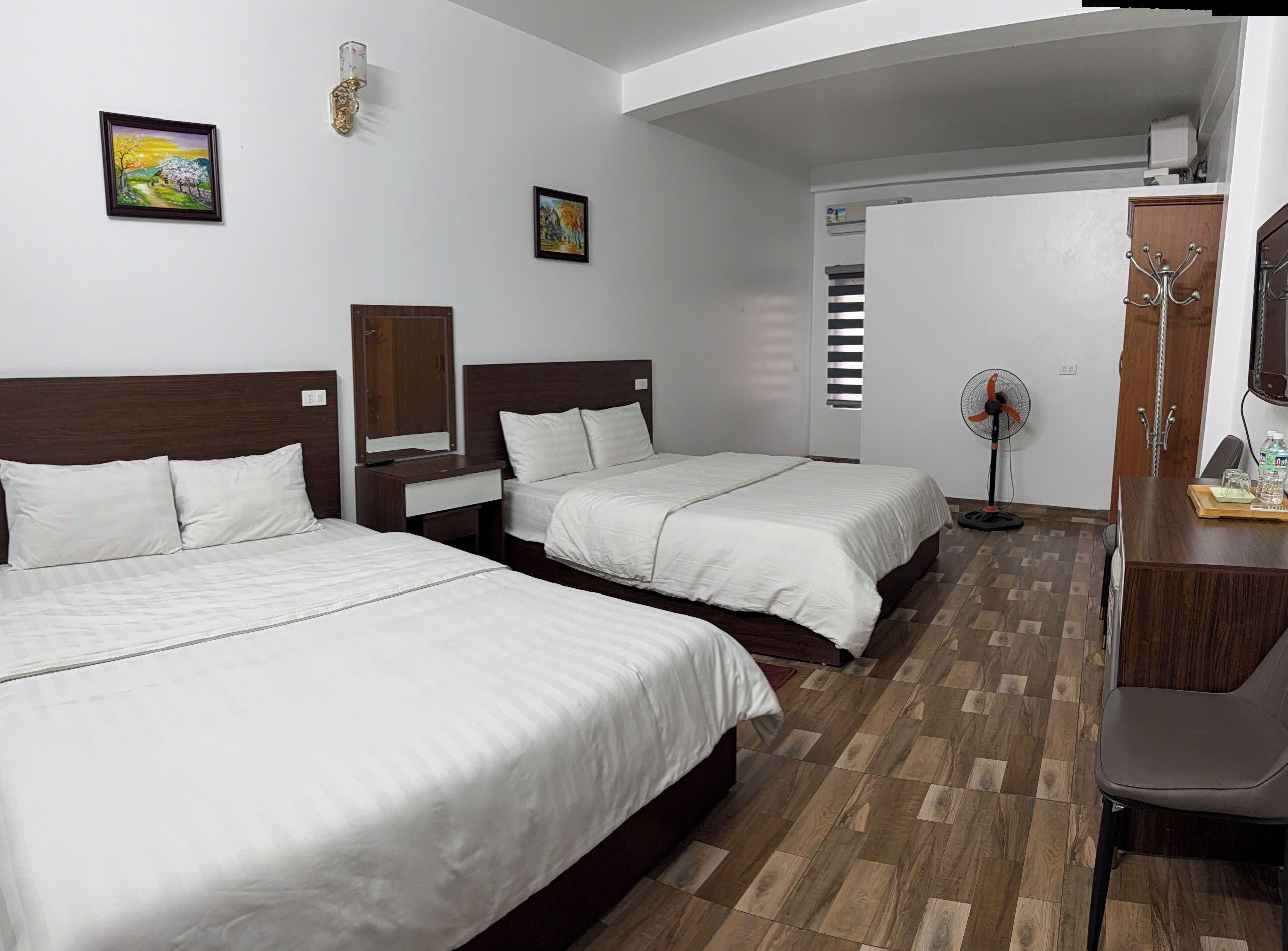 A hotel room with two single beds, a wooden desk, a chair, a TV, and a fan.