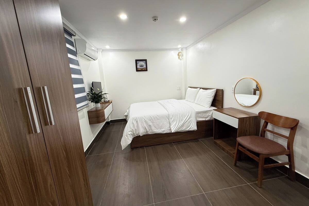 A bedroom with a bed, a desk, a chair, a wardrobe, a bedside table with a plant, and a mirror on the wall.