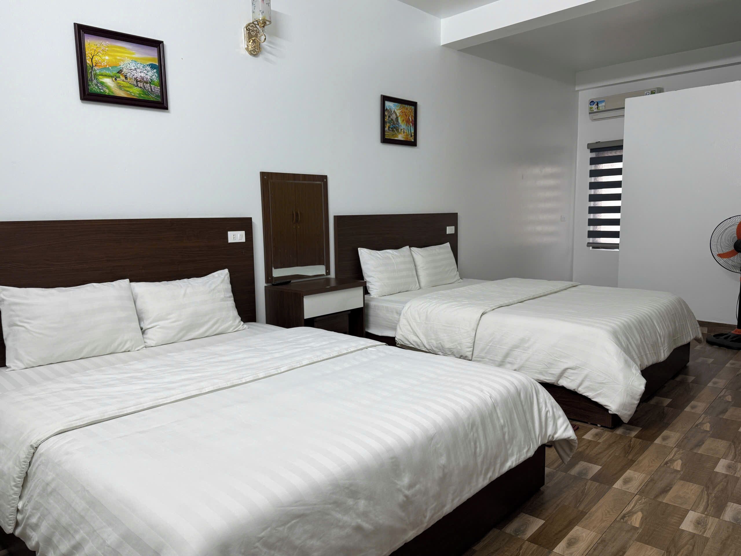 Habitación de hotel con dos camas individuales, cabecero de madera, mesita de noche, espejo y un ventilador de pie.