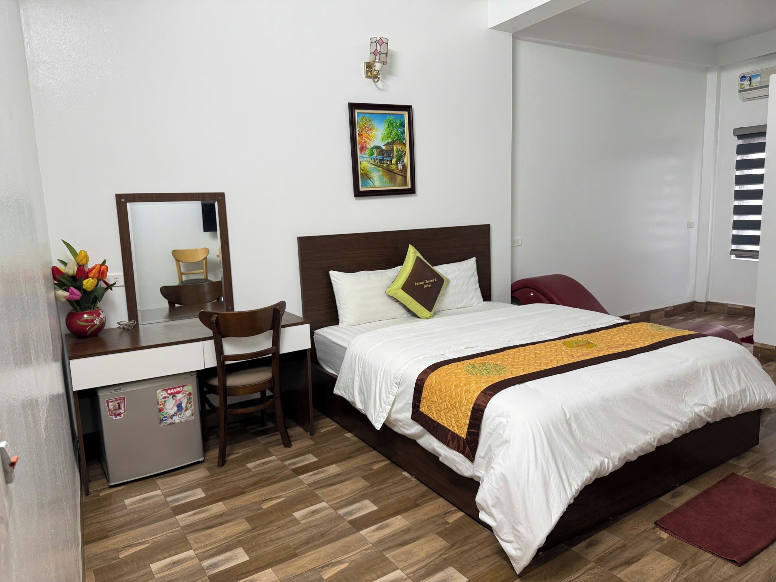 Habitación de hotel con cama, escritorio con silla, refrigerador pequeño, espejo, un cuadro en la pared y ventana con persianas.