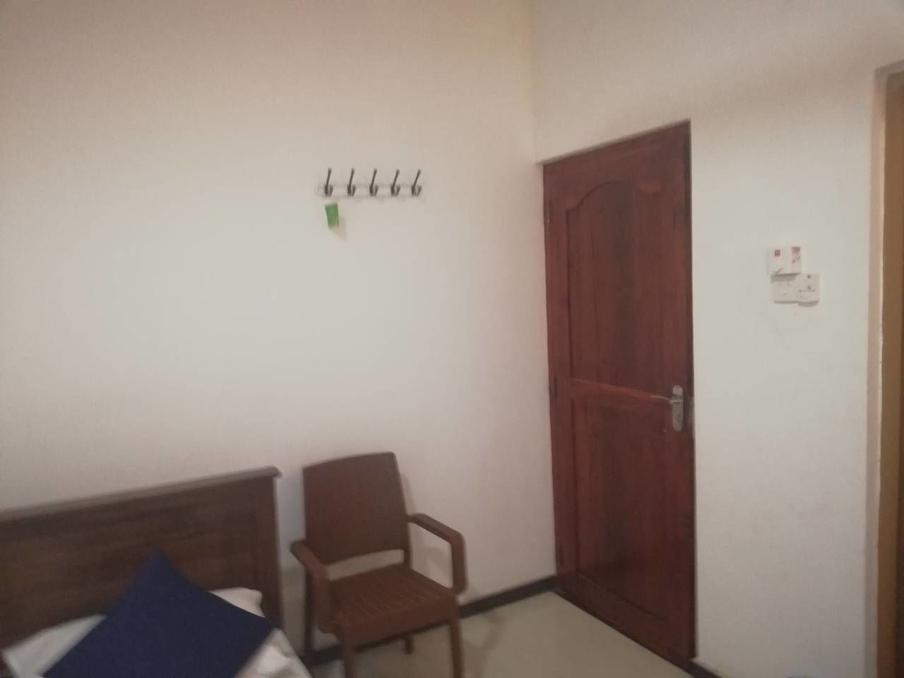 Una habitación con una cama de madera, una silla, una puerta de madera y un gancho en la pared con una etiqueta verde.