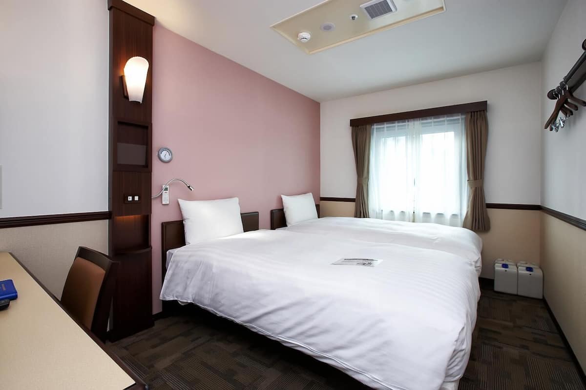 A hotel room with a large bed, a desk, a chair, a bedside table, a clock, a window with curtains, and a wall-mounted heater.
