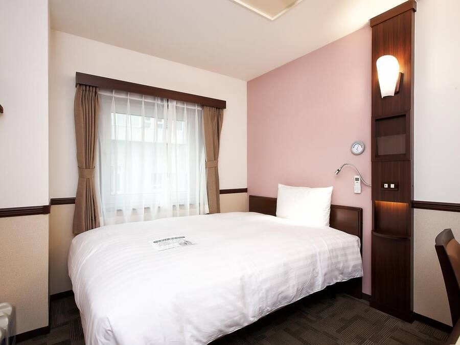 Una habitación de hotel con una cama grande, una mesita de noche, una lámpara de pared y una ventana con cortinas.