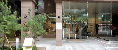 A hotel entrance with a glass facade, a sign reading "toyoko-inn.com", and a small outdoor area with plants.