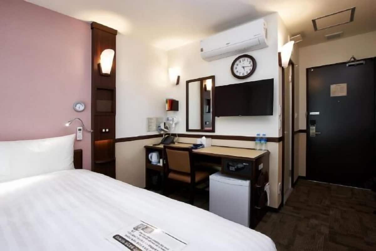 A hotel room with a bed, a desk, a chair, a small refrigerator, a clock, and a door.