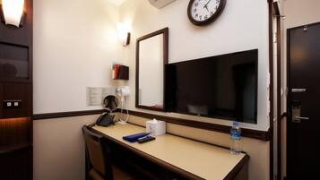 A hotel room with a desk, chair, clock, TV, and a minibar.