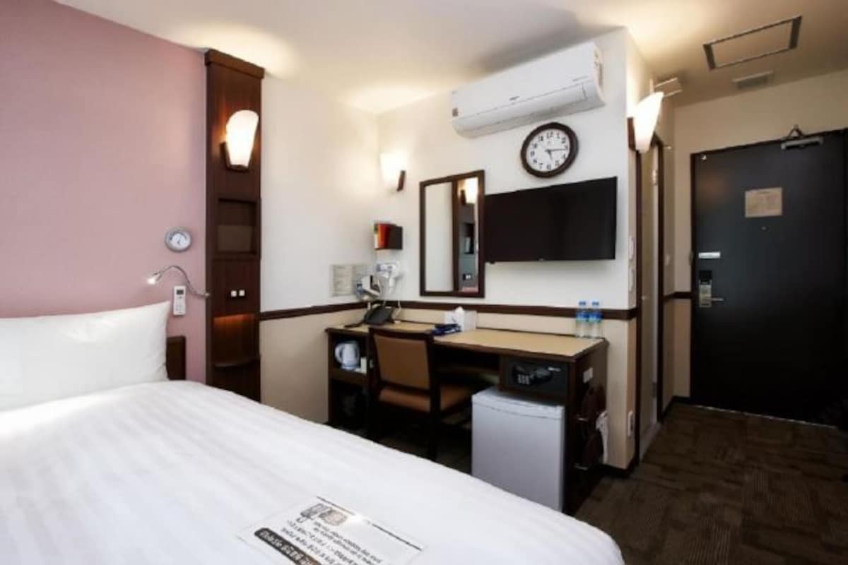 Une chambre d’hôtel avec un lit, un bureau, une chaise, un petit réfrigérateur, une horloge et une porte.