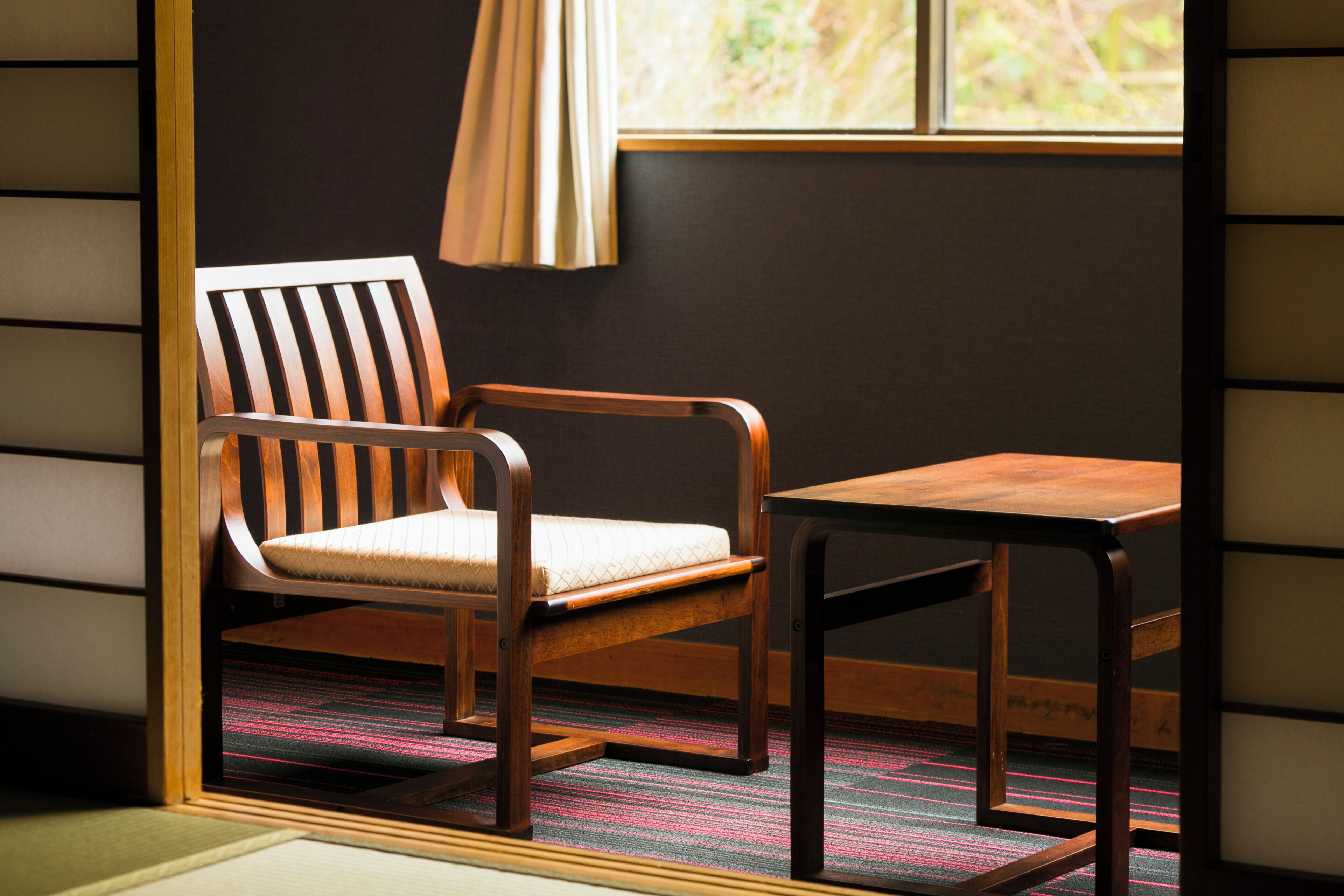A wooden chair with a cushioned seat and backrest, a matching wooden table, and a window with curtains.