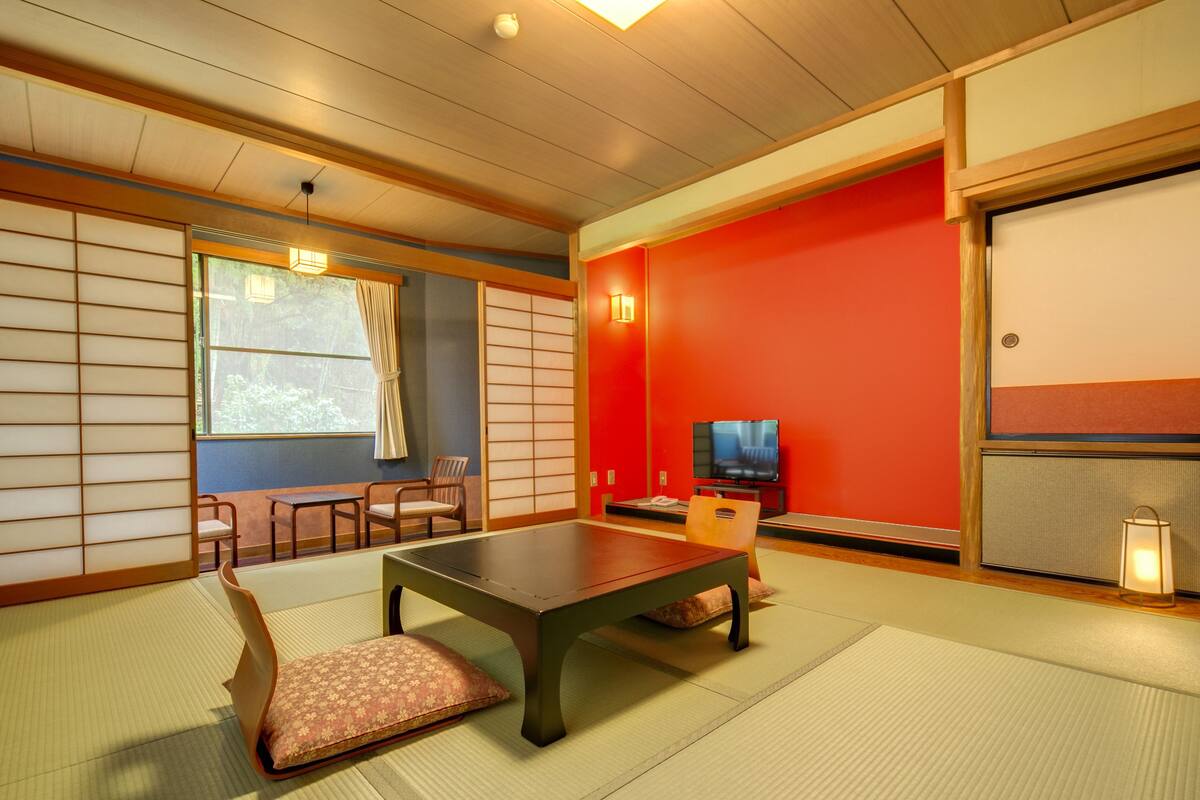 Ein traditioneller japanischer Raum mit Tatami-Boden, einem niedrigen Tisch, Kissen, einem Fernseher und Schiebetüren.