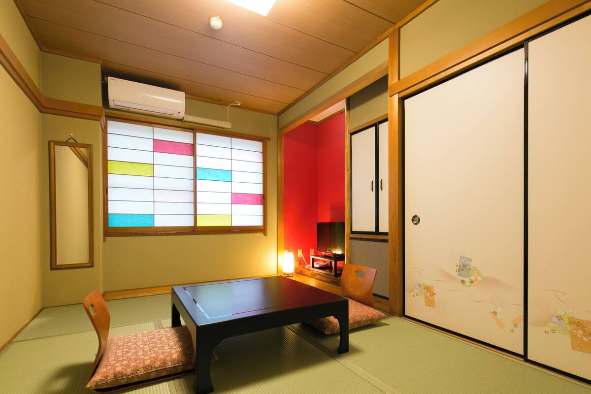 Ein traditioneller japanischer Raum mit Tatami-Böden, einem niedrigen Tisch, einem gepolsterten Sessel, einer Schiebetür und einem Fenster mit farbigem Papier.