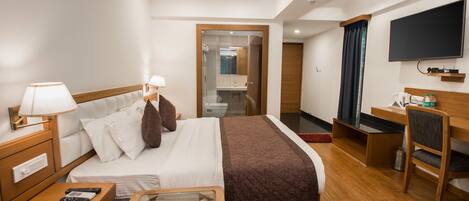 A hotel room with a bed, bedside tables, a desk, a chair, a TV, and a bathroom visible through a glass door.