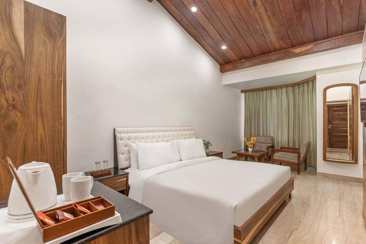 A hotel room with a large bed, wooden headboard, bedside tables with lamps, a small table with a vase of flowers, and a mirror on the wall.
