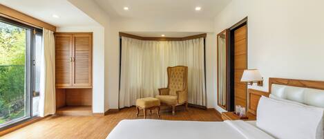 Une chambre d’hôtel avec un grand lit, un plancher en bois, une chaise, un bureau avec une lampe et une vue sur la verdure par la fenêtre.