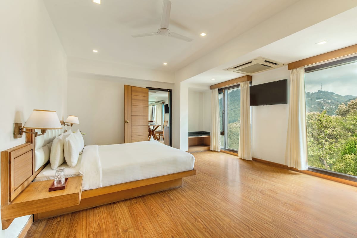 A spacious bedroom with a large bed, wooden headboard, bedside lamps, a flat-screen TV, and a view of a mountainous landscape through large windows.