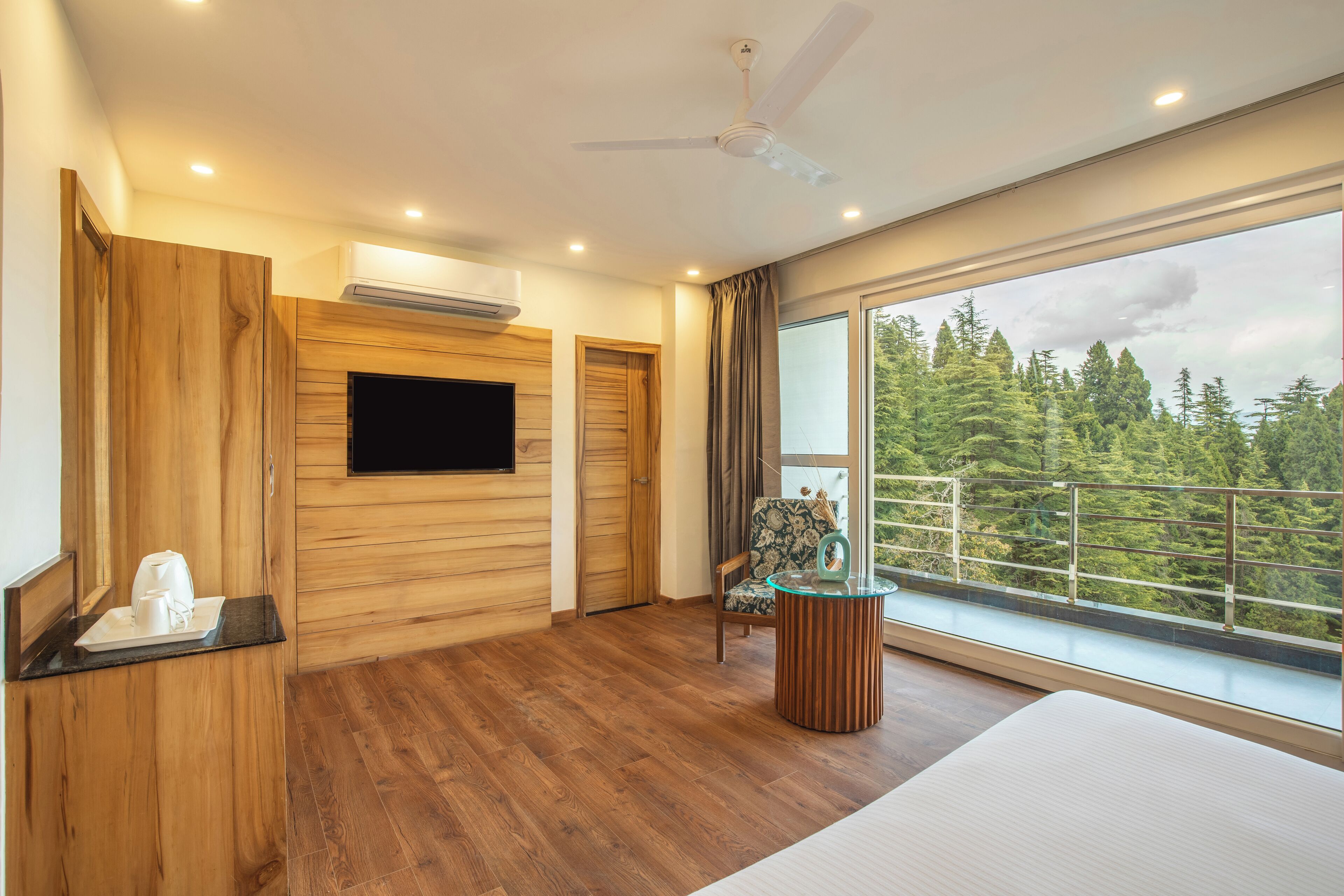 A modern hotel room with a wooden floor, a flat-screen TV mounted on the wall, a large window with a view of trees, and a ceiling fan.