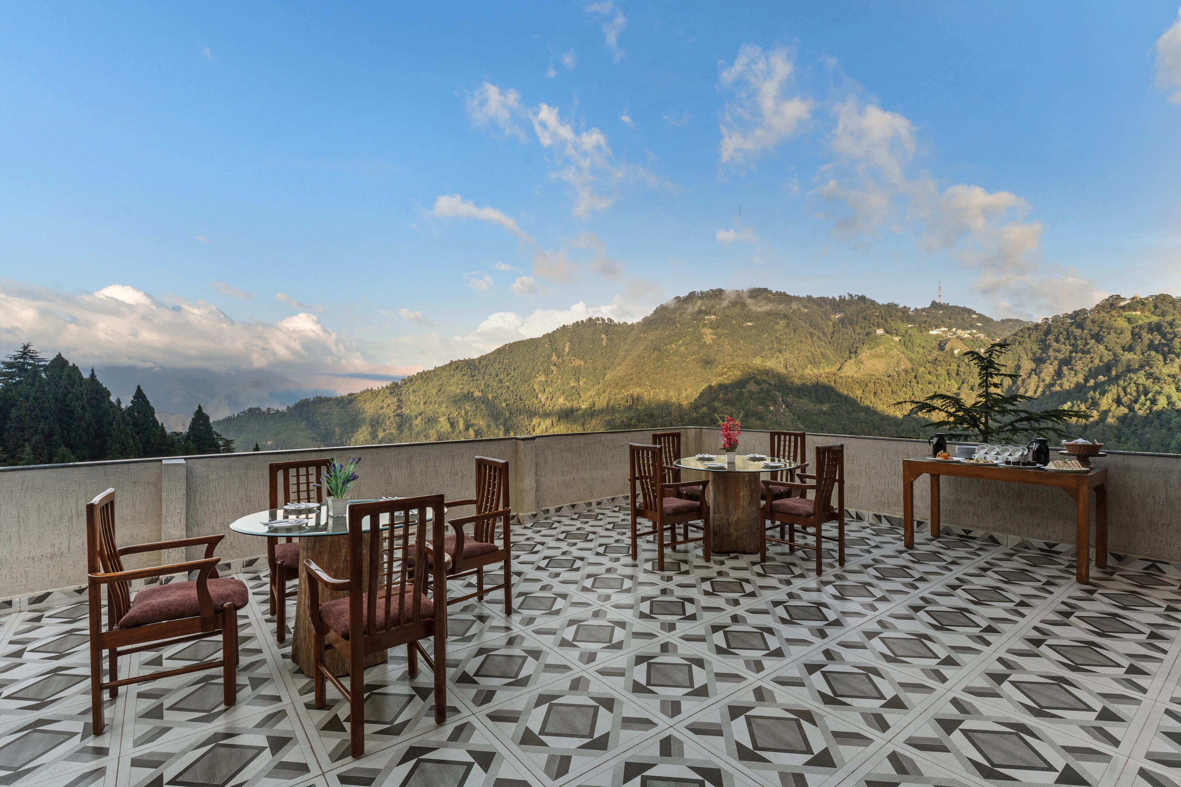 A terrace with a geometric patterned floor, wooden furniture, and a view of a forested mountain range.
