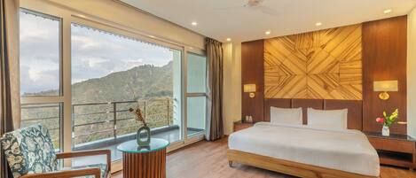 Quarto de hotel com uma cama grande, uma cadeira, uma mesinha e vista para uma paisagem montanhosa através de amplas janelas.