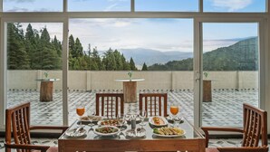 Mesa de jantar com pratos de comida e bebidas, com uma paisagem montanhosa ao fundo, visível através de amplas janelas.