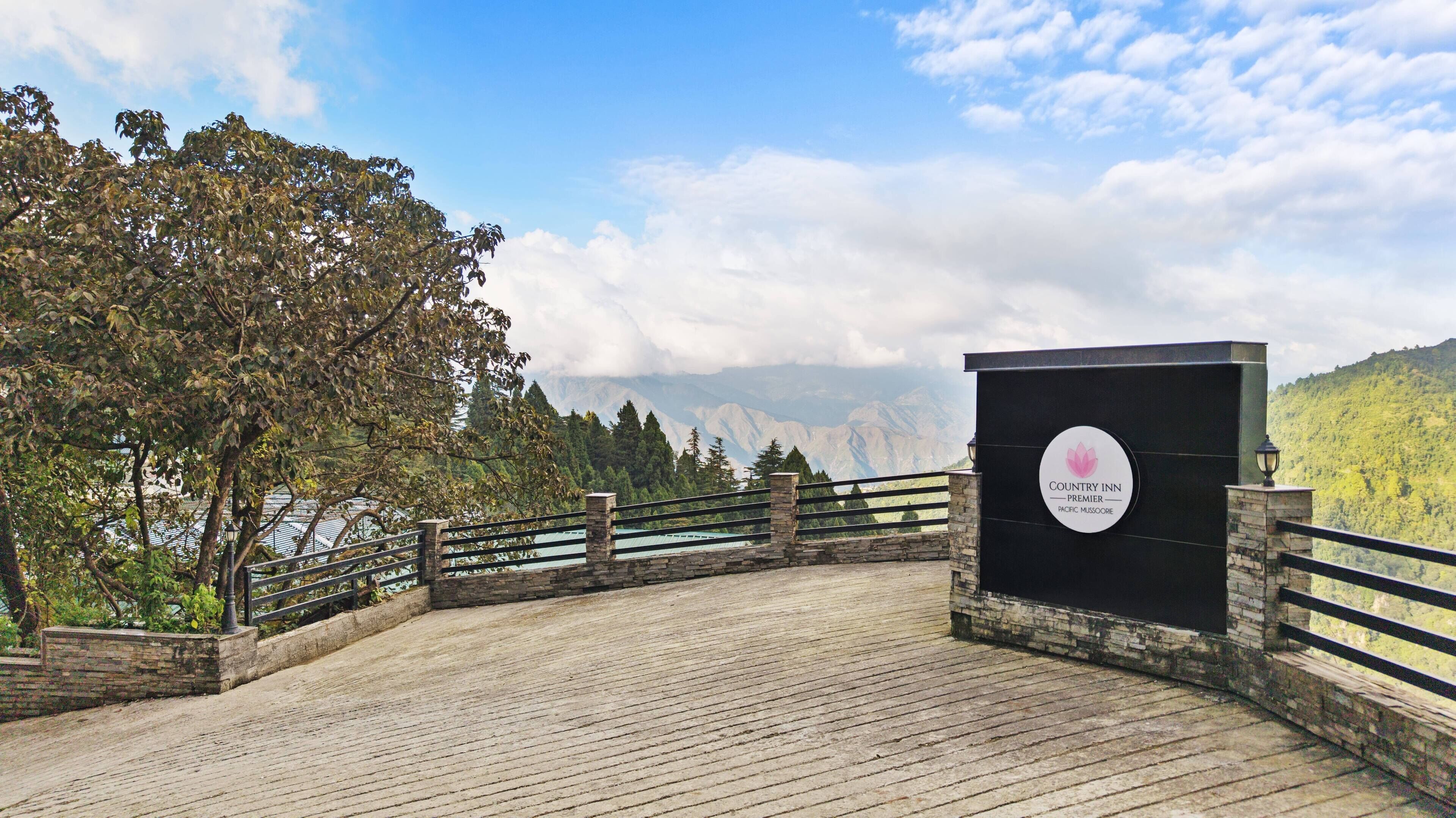 Una terraza de madera con baranda de piedra, un cartel de Country Inn Premier Pacific Mussoorie, y un paisaje montañoso al fondo.
