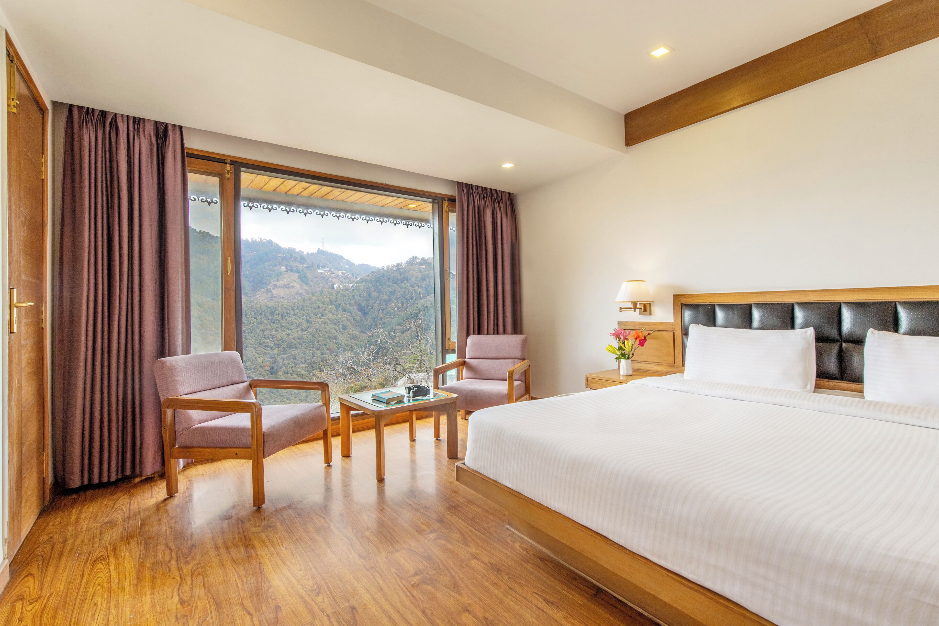 A hotel room with a large bed, two armchairs, a small table, and a view of a mountainous landscape through large windows.