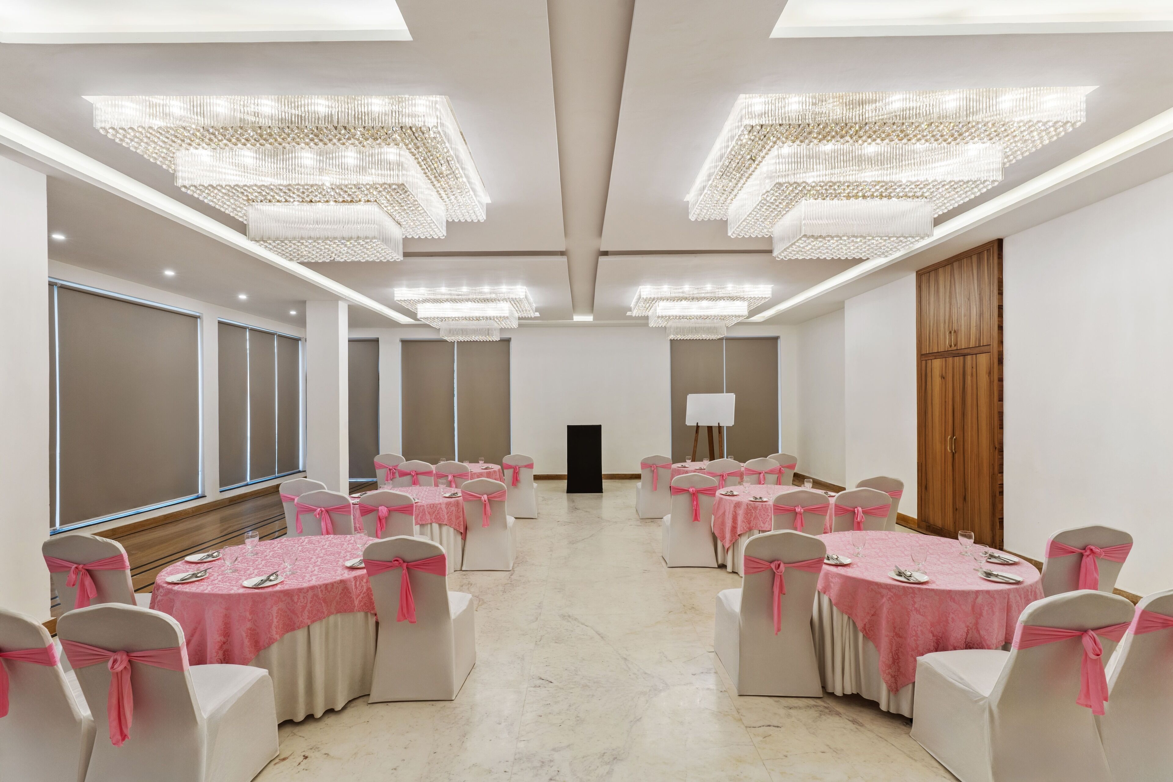 Un salón de conferencias con mesas redondas, sillas blancas adornadas con cintas rosas y grandes candelabros.