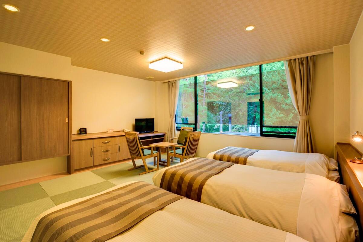 A hotel room with two beds, a desk with a chair, a television, and large windows with curtains.