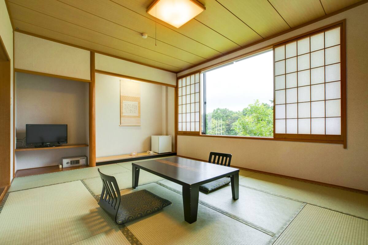 A traditional Japanese room with tatami flooring, a low table, chairs, a television, and a large window with shoji screens.