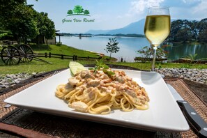 A plate of pasta with a glass of white wine, set against a scenic lakeside view with a wooden cart and a fence. - Green View Resort (Vang Vieng)