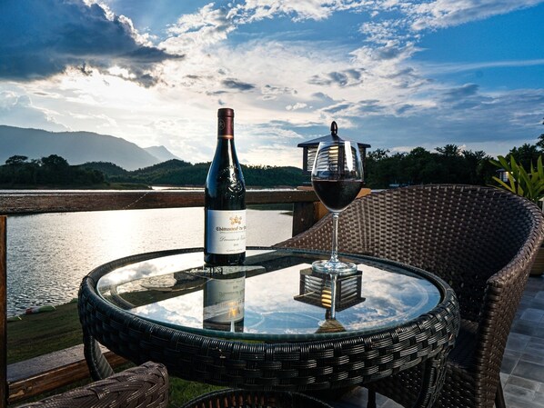 A bottle of wine and a glass on a glass table with a wicker chair beside it, overlooking a body of water and mountains. - Green View Resort (Vang Vieng)