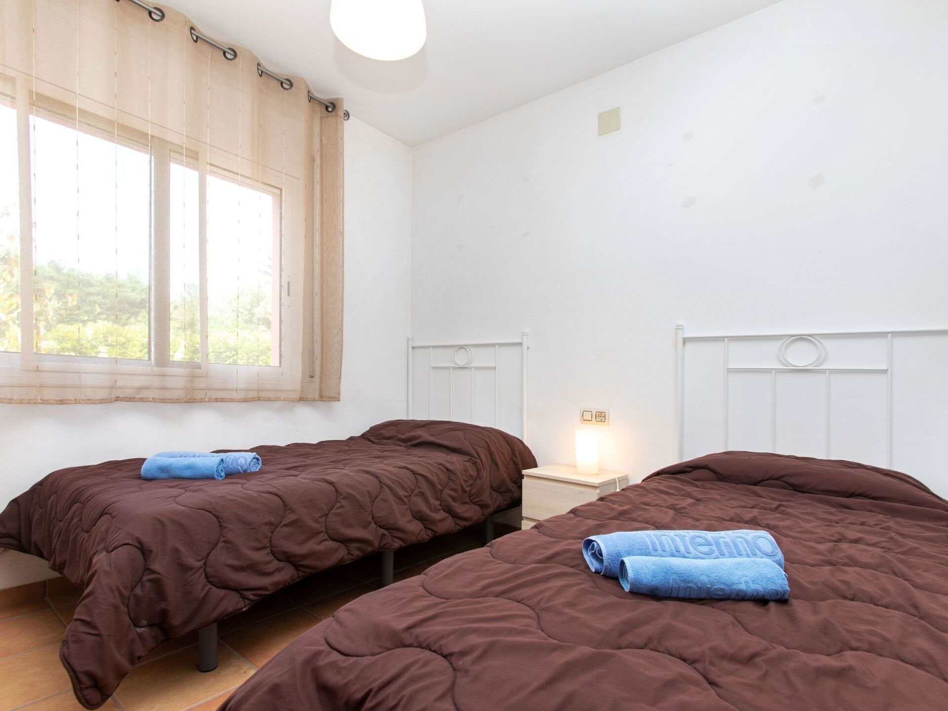 Une chambre avec deux lits, une fenêtre avec des rideaux, une table de chevet avec une lampe, et une vue sur la verdure à l’extérieur.