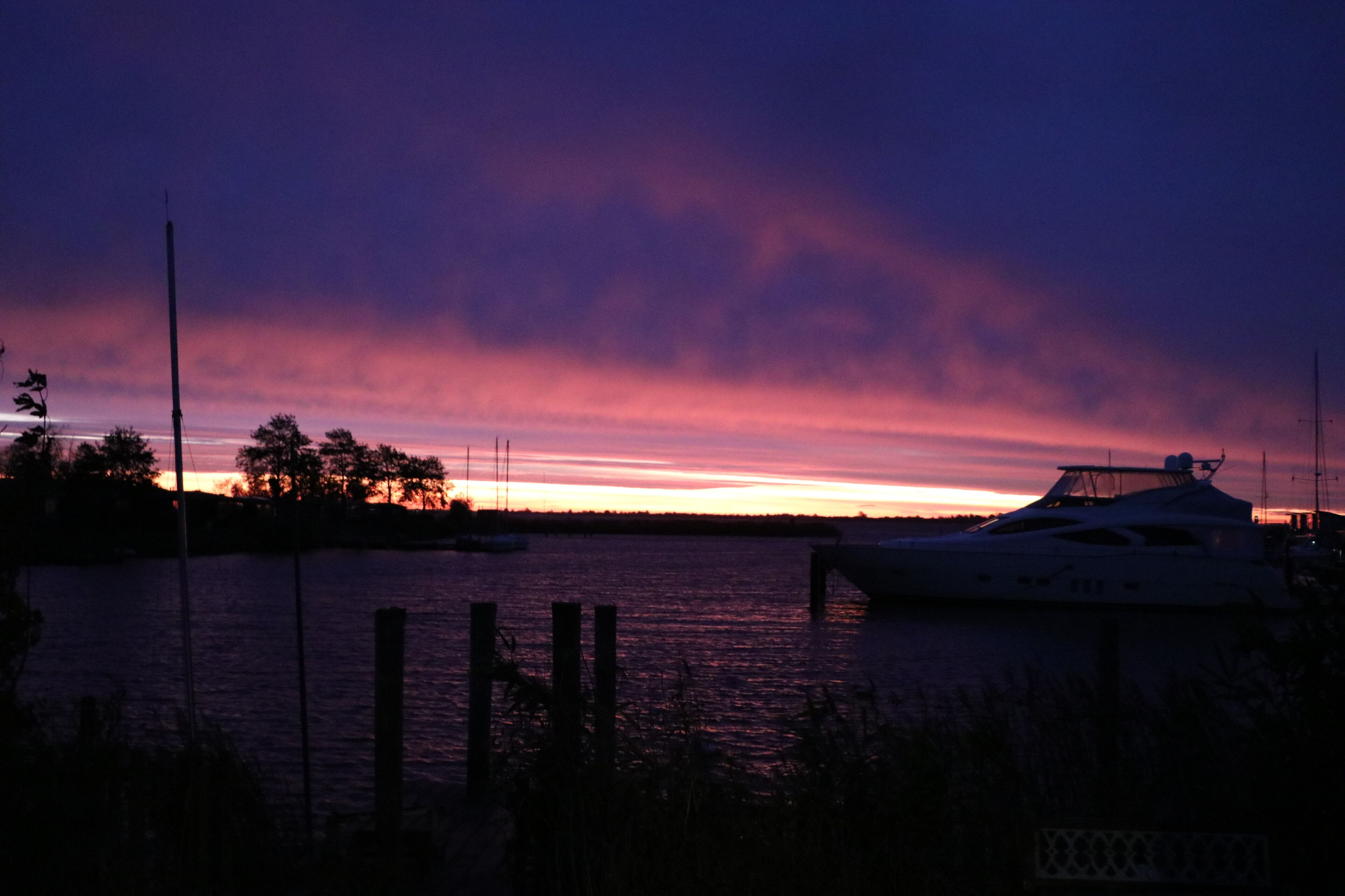 Eine Uferlandschaft mit einer am Steg vertäuten Yacht, umgeben von ruhigem Wasser und einem Sonnenuntergangshimmel.