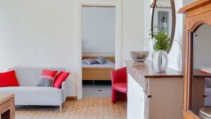En hyggelig stue med en grå sofa, røde puder, en rød lænestol og et træbord. En seng med mønstrede puder ses i baggrunden.