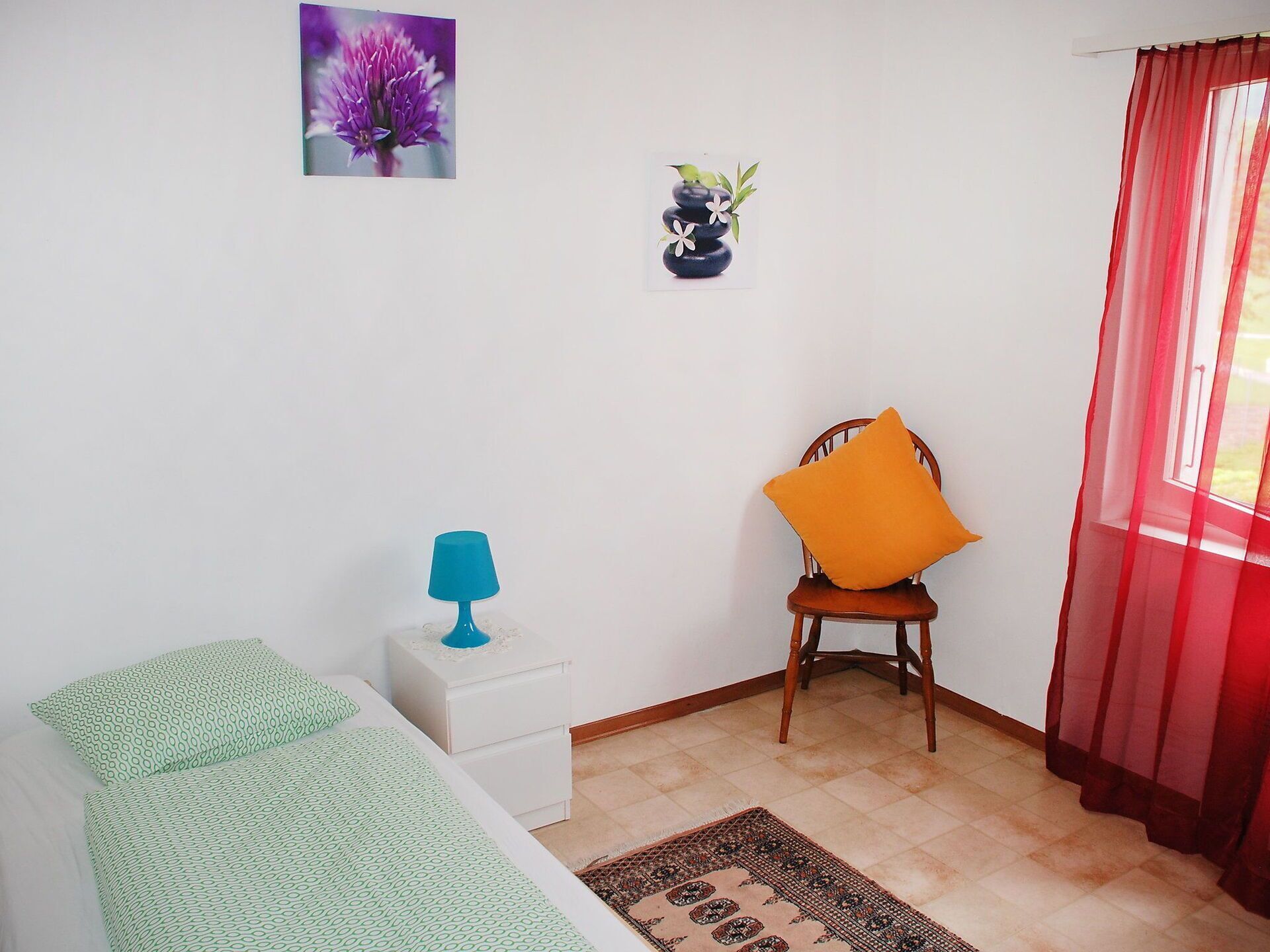 Een compacte, verzorgde kamer met een bed, een stoel met oranje kussen, een tafeltje met lamp, een wandvaas en een raam met rode gordijnen.